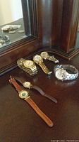Six watches arranged on a dark wooden surface near a mirror, showing a mix of metal and leather strap watches
