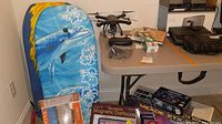 The lot items arranged including a blue dolphin bodyboard, Holy Stone drone and accessories, books, poker chips, reversible dartboard, and other items on table and floor.
