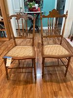 Front view of two small scale antique wooden chairs placed side by side on a wooden floor near a doorway. One chair has a more ornate backrest carved in the shape of a crown (Queen), the other has a simpler carved back (King). Both have upholstered seats.
