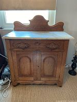 Vintage wooden wash stand with carved front doors, single drawer, and decorative backsplash, standing on carpeted floor near a window. Electrical cords visible beside it.