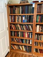 Front view of a tall oak wood bookcase filled with various hardcover books on five shelves, showing natural wood grain and bezel shelf edges.