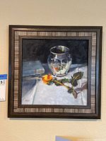 Framed still life painting of a glass vase with a yellow-orange rose on a white cloth with a partially visible book in the background.