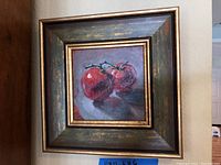 Frontal view of the small framed oil painting of two red tomatoes on a surface with visible signature.