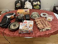 Table display showing various bakeware including pans, trays, a boxed cookie press, cupcake pan, cooling rack, and Salton brand electric grill sandwich/waffle maker with cord.