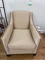 Front view of beige upholstered armchair with nailhead trim and wooden legs.