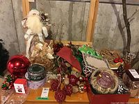 Full lot of Christmas decorations on wooden table with cellar wall backdrop, showing Santa, angel, candle holders, tins, ribbons, ornaments.