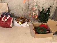 Photo showing green artificial Christmas trees, present bags in red and blue, and cardboard box containing wreaths and decorative materials.