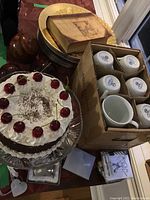 Image showing faux cake, gold and silver chargers, coffee mugs in box, decorative balls, and invitation cards on a table.