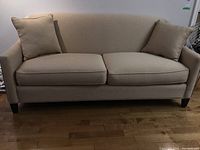 Front view of beige upholstered couch with 2 cushions, wooden legs visible, on hardwood flooring.
