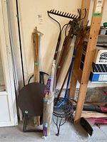 Full view of garden tools bundled together including rakes, hoes, bamboo stakes, two small round metal tables leaning against wall, metal plant hook, circular metal plant supports, and an open burlap bag on the ground.