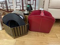 Photo showing three women's hats: a blue wide-brimmed hat with striped ribbon, a plaid cap-style hat in a striped box, and a red leather magazine basket beside the box.