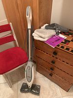 Front view of Shark Pro Steam Pocket Mop and accessories on a wooden chest next to a red padded chair in a tiled floor room.