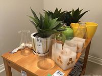 Photo of various home decor items on a wooden table including faux succulent plants in ceramic planters, yellow ceramic pots, green bird figurines, clear glass hurricane candle holders, and square block candles with geometric patterns.