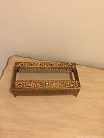 Ornate gold filigree tissue box cover with pink stones, rectangular shape, elevated on small legs.