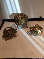 Set of three brass tone oval Art Nouveau style jewelry boxes with embossed swirls and flowers, placed on a white surface, showing overall condition and size comparison.