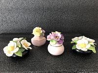 Four bone china flower ornaments: two black vases with white flowers and two pink vases with gold strip and multicolored flowers, arranged on a black surface.