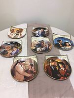 Photograph of 7 Gone with the Wind collector plates arranged on a table with certificates of authenticity beneath them. Plates depict colorful scenes from the movie with gold rims, no visible damage.