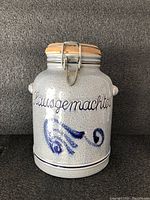 Front view of stoneware kitchen storage pot with blue 'Hausgemachtes' text and decorative design, wooden lid and metal clasp locking mechanism.