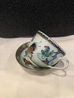 Side view of hand-painted porcelain teacup resting on saucer, showing detailed rooster and floral pattern with gold trim.