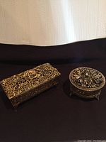Set of two brass jewelry boxes, one rectangular and one round, shown closed on black fabric background