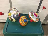 Three vintage tin spinning top toys shown on a green surface, each with a plastic spinning handle and metal spiral rod, highlighting colorful animal and cartoon designs.