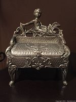 Front view of vintage cast metal pewter jewelry box with cherub figure on back edge, showing detailed embossed design and front feet.