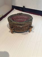 Closed view of oval brass filigree jewelry box with clear glass lid and cherub feet.