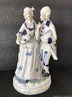 Porcelain Victorian-style figurine of a romantic couple, white base with blue painted details including floral motifs. Man and woman in period costume standing close, woman holding a fan.