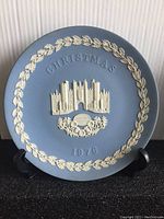 Front view of Wedgwood commemorative plate with raised cream-colored relief of Hampton Court castle against a matte blue background, surrounded by a decorative laurel wreath and inscription "Christmas 1976." Plate rests against a black stand (stand not included in sale).