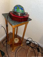Full view of the tall wooden corner table with green marble-look top and lower shelf with boxes, lamp on top.