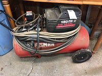 Full side view of red cylindrical tank air compressor with black top housing. Two black wheels for portability. Hoses coiled on unit.