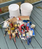 Top-down view of the lot with plastic buckets, various paint brushes, paint rollers, adhesive tubes, and a scrubbing brush arranged on a deck.