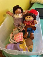 Two Cabbage Patch Kids dolls lying in clear plastic tote with spare clothes.