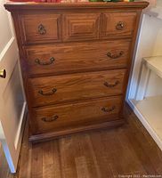 Front view of the wooden chest of drawers with metal handles, showing the top three smaller drawers and the larger bottom drawers, placed on a wooden floor next to a white shelving unit.