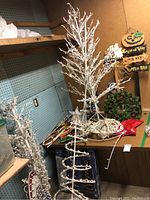 White artificial Christmas trees with attached lights, coiled Christmas light strings, wreath, and Halloween signs visible in storage setting.