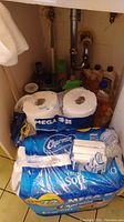 Photo of under-sink cabinet shelves with multiple packs of Charmin toilet paper, both mega and ultra soft, and small Kleenex tissue packs