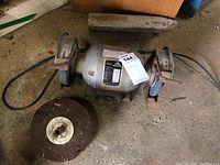 Full view of Delta bench grinder showing both grinding wheels and power cord on concrete floor with additional grinding stones nearby.