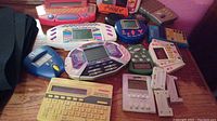 Lot of 10 electronic handheld games including Wheel of Fortune, Stop!, Who Wants to Be a Millionaire, poker, and other games arranged on a wooden surface, showing colorful plastic casing and many buttons.
