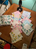 Overview of multiple floral handkerchiefs in pastel colors spread on a wooden surface under lamp light showing various floral patterns and scalloped edges.
