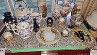 Photo showing a collection of ceramic and porcelain decorative items including vases, plates, a teapot, and trinket boxes with floral designs in various styles and colors.