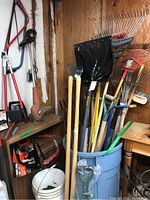 Photo showing assorted rakes, shovels, brooms, snow shovels stored in bins and against wall in a workshop