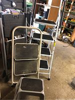 View of multiple step stools showing tall white four-step stool with black treads, smaller 2-step and 3-step stools with silver or gray frames and black steps.
