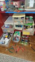Photo showing collection of plastic storage containers, multiple kinds of pliers spread on floor, assorted craft materials packed in drawers