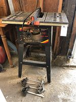 Front view of Menards 10" table saw on stand with two miter gauges on floor.