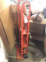 Side view of folded metal utility dolly leaning against a wall with a white label hanging from the handle.