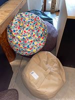 Four bean bags stacked and arranged near stairs and furniture, colorful triangle patterned bean bag on top, beige bean bag with handle front right, purple and dark green bean bags partially visible in the back.