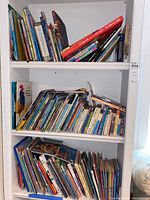 Three white shelves densely packed with children's books arranged upright and stacked.