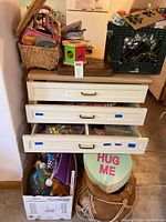 Three white drawers, partially open, filled with various crafting and small items, a box of stuffed toys underneath, wicker baskets, and a 'HUG ME' sign visible.