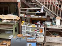 Photo of a white and wooden multi-tier shelf filled with small items like paint tubes, containers, and boxes. Also visible is a green wooden workbench with drawers and scattered tools and hardware.
