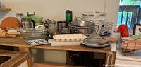 Wide view of assorted kitchenware on a wooden surface showing pots, pans, bowls, egg holder, and kitchen utensils.